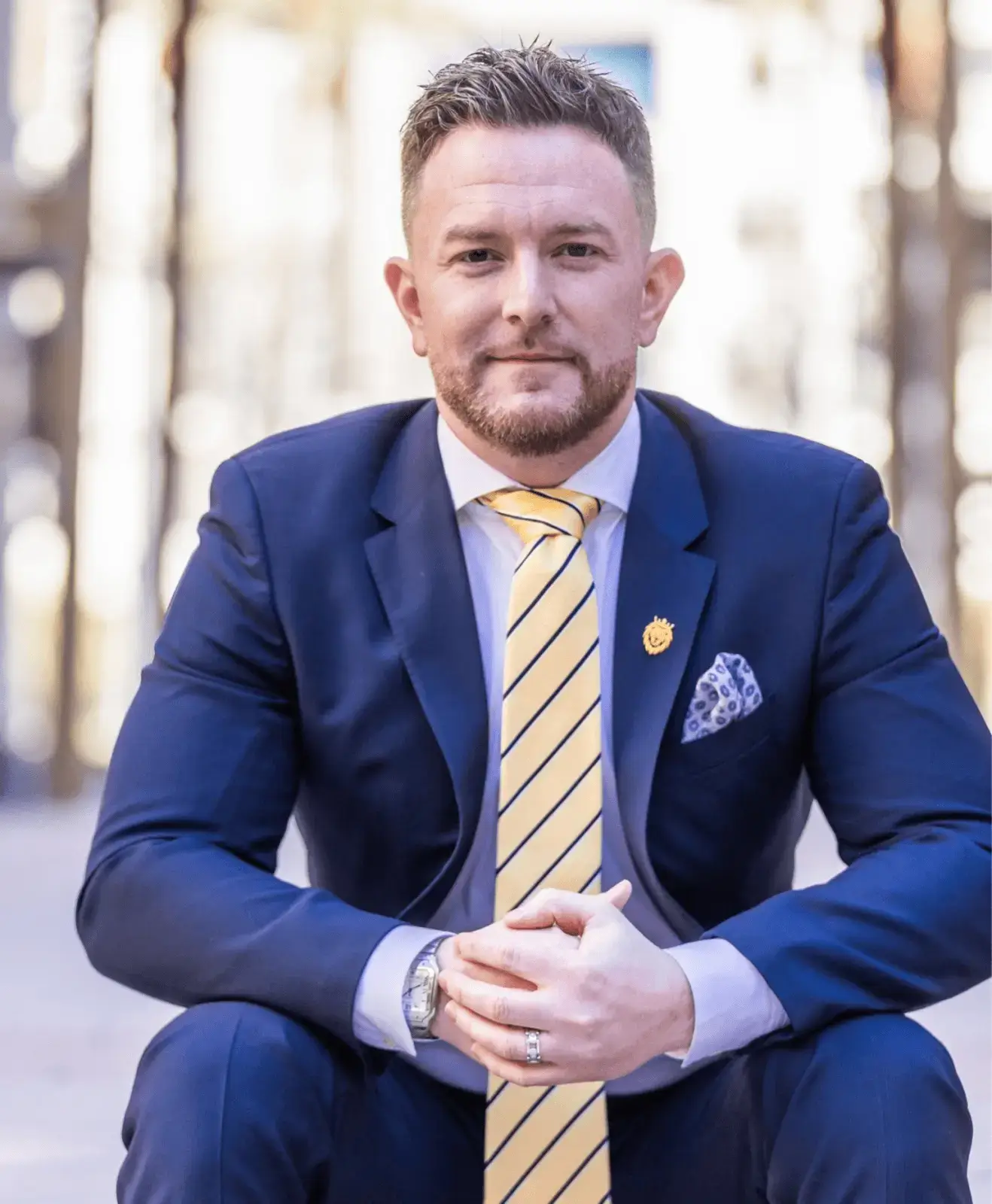 A man in a navy suit with a yellow striped tie and pocket square sits outdoors, looking at the camera with hands clasped.