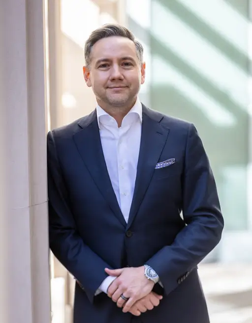 A man in a dark suit and white shirt stands indoors, leaning against a wall, with hands clasped in front of him. Light and shadows fall across the background.